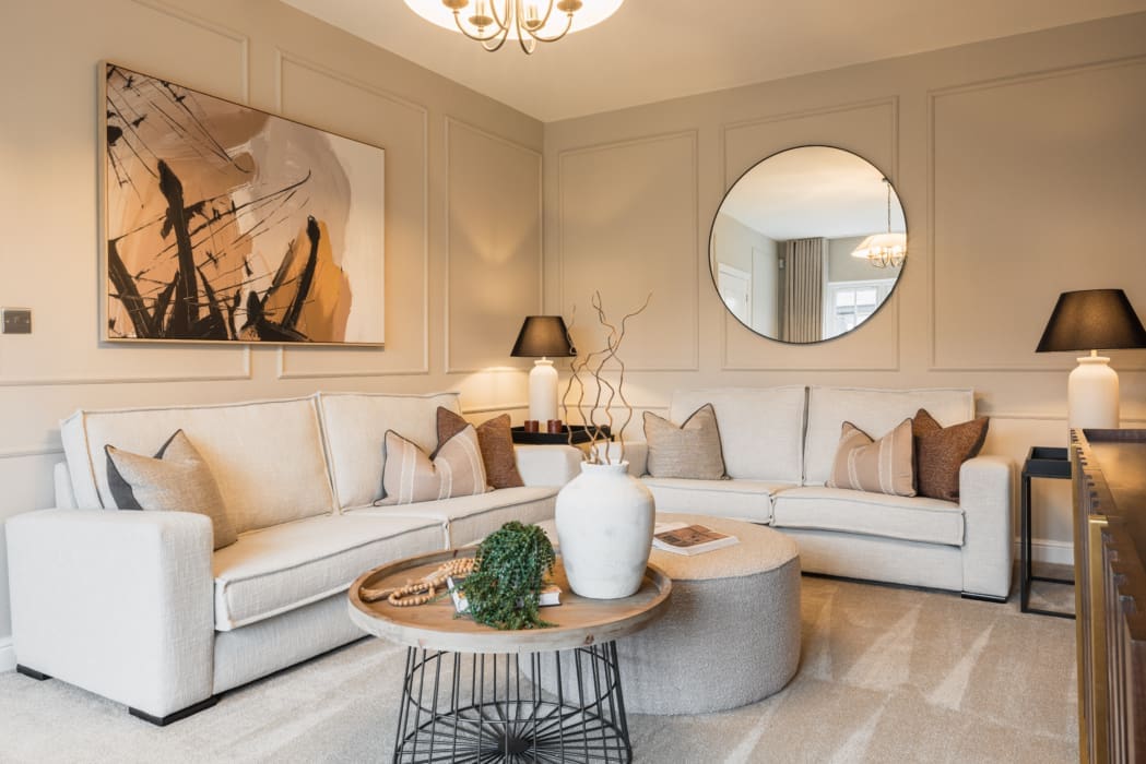 A stylish living room in a new build home by Anwyl Homes, featuring beige sofas adorned with cushions, a round mirror, modern artwork, and accent tables with decorative lamps.