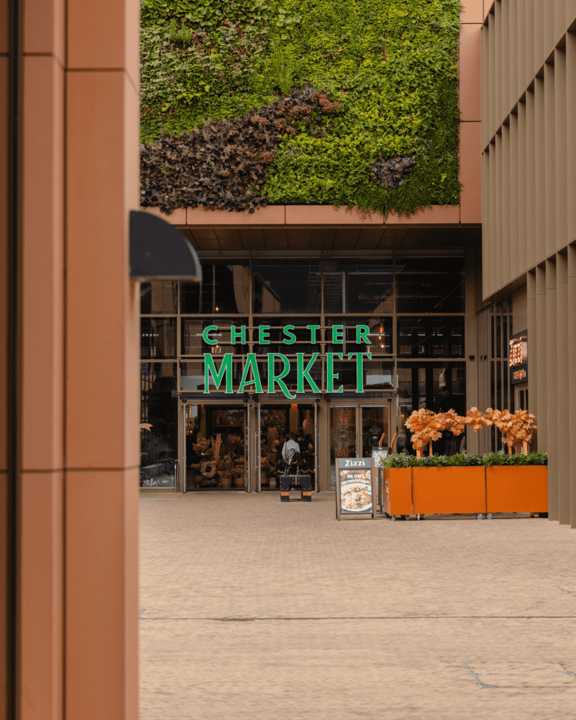 A modern entrance to Chester Market is framed by vertical greenery. Orange planters with foliage adorn the pathway, and a neon sign welcomes visitors to the bustling marketplace.