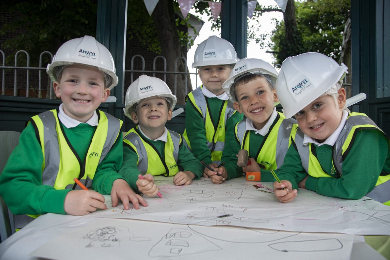 Five smiling children wearing hard hats and high-visibility vests are drawing on a large paper sheet. They sit around an outdoor table, suggesting a playful, learning environment.