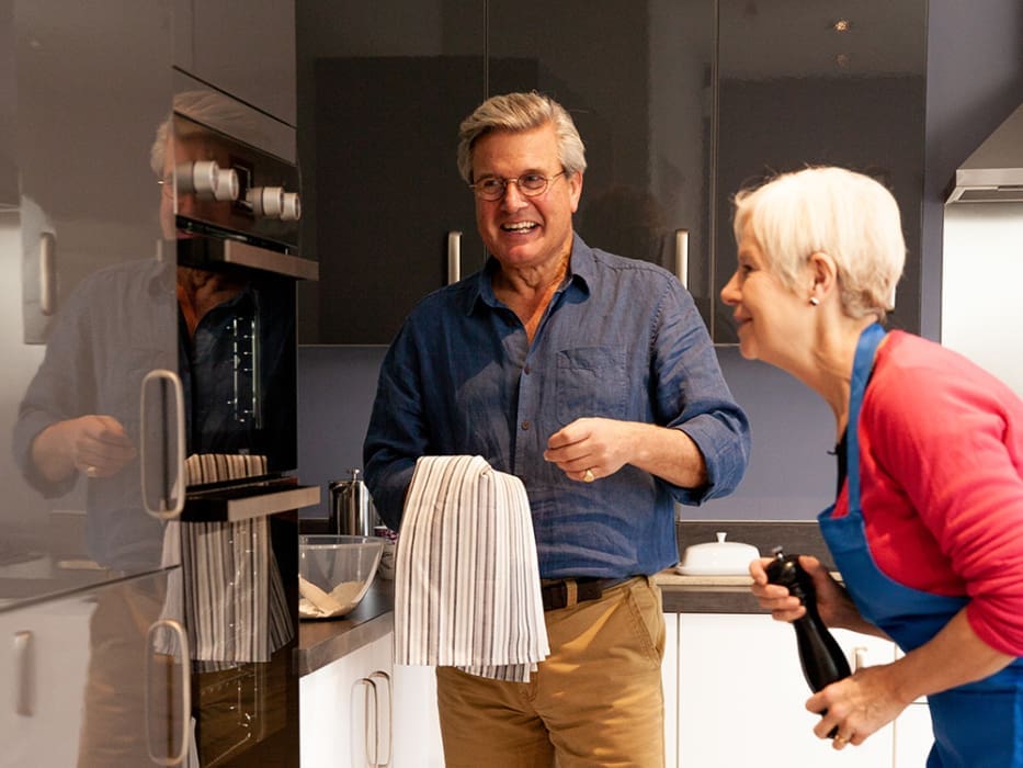 A cheerful man and woman are cooking together in a modern kitchen, with dark cabinets and a sleek oven. The woman is holding a pepper grinder, while the man uses a towel.
